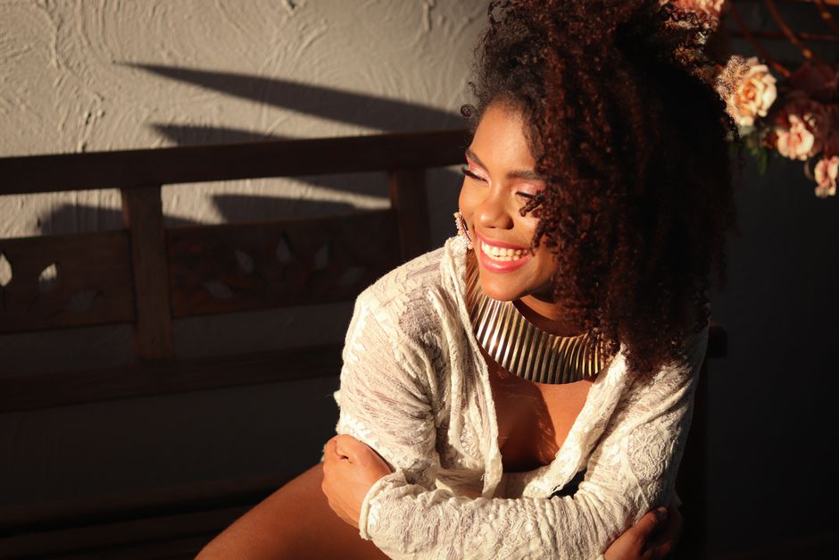 A joyful woman with an afro smiles warmly in a sunlit room, wearing a stylish necklace.