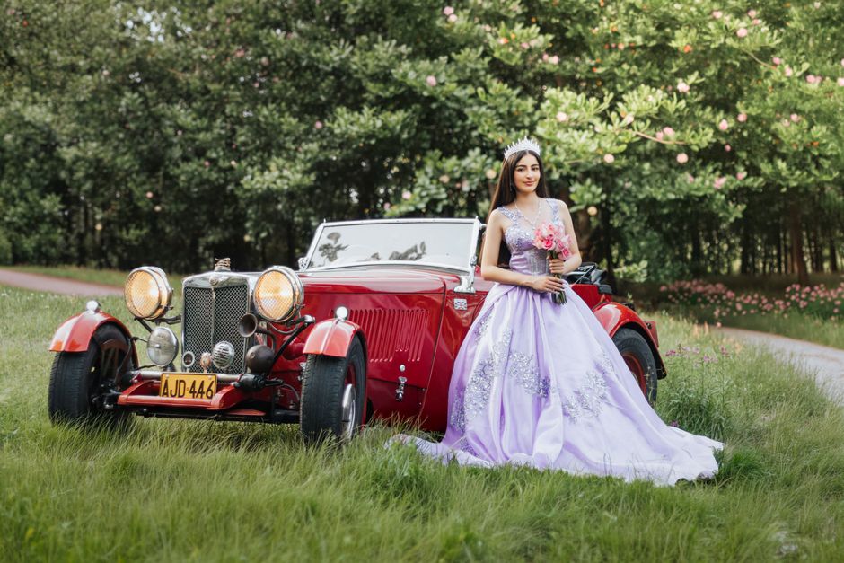 A young woman in a lavender dress poses beside a vintage red car in a lush outdoor setting in La Dorada, Colombia.