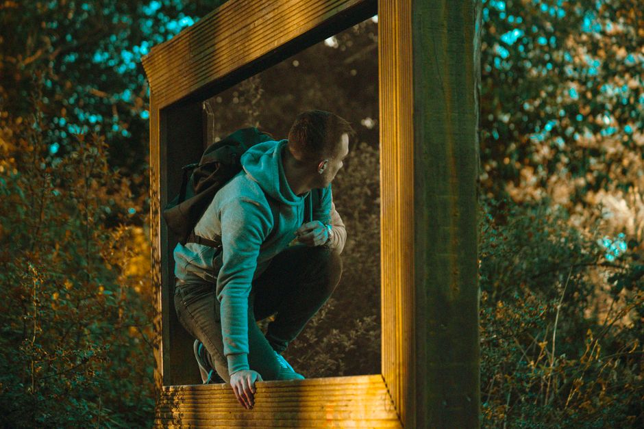 A man in a gray hoodie kneels amidst vibrant foliage, peering through a rustic wooden frame.