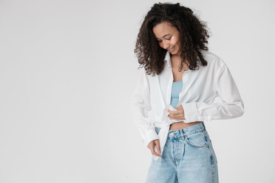 Young woman in a casual outfit smiling and adjusting her denim jeans against a neutral background.
