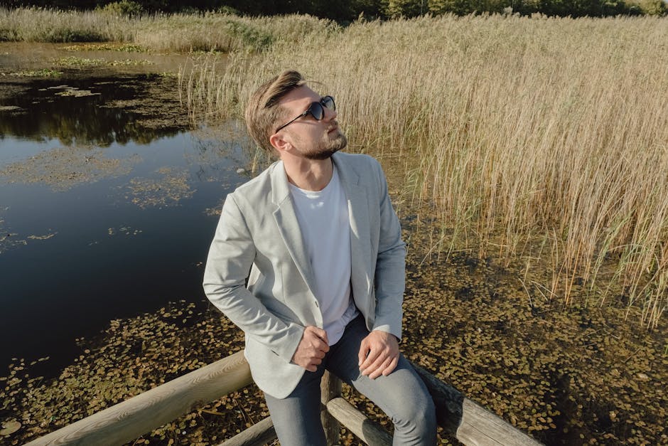A man in sunglasses and a blazer enjoys a sunny day by a peaceful lakeside.