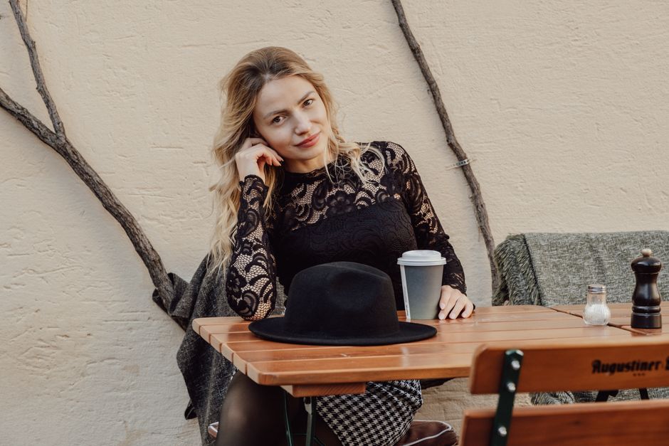 Woman in stylish lace top sitting at a wooden table outdoors with coffee and hat.