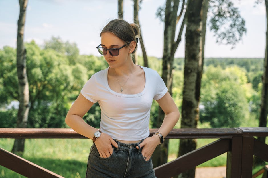 A woman wearing sunglasses and a white blouse stands confidently in a sunlit park setting.