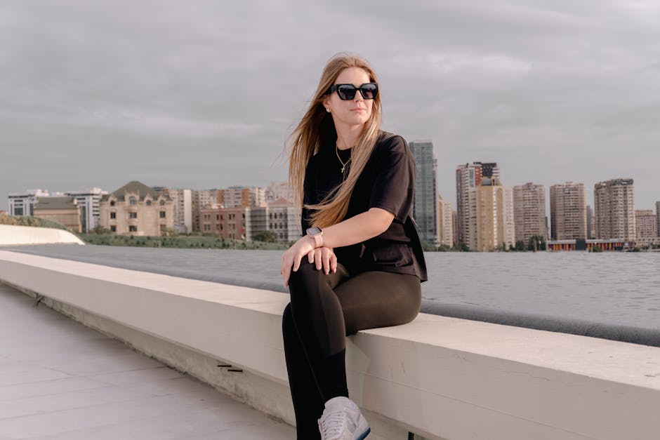 Stylish woman sitting by a waterfront with a city skyline in the background. Casual and urban scene.