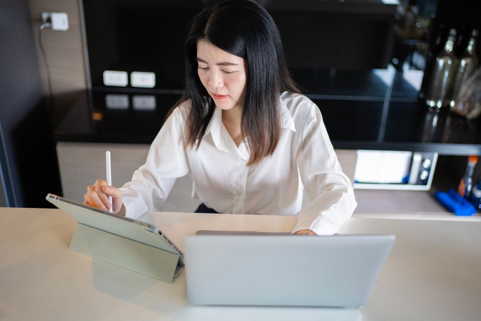Asian woman multitasking with a laptop and tablet in a modern home office.