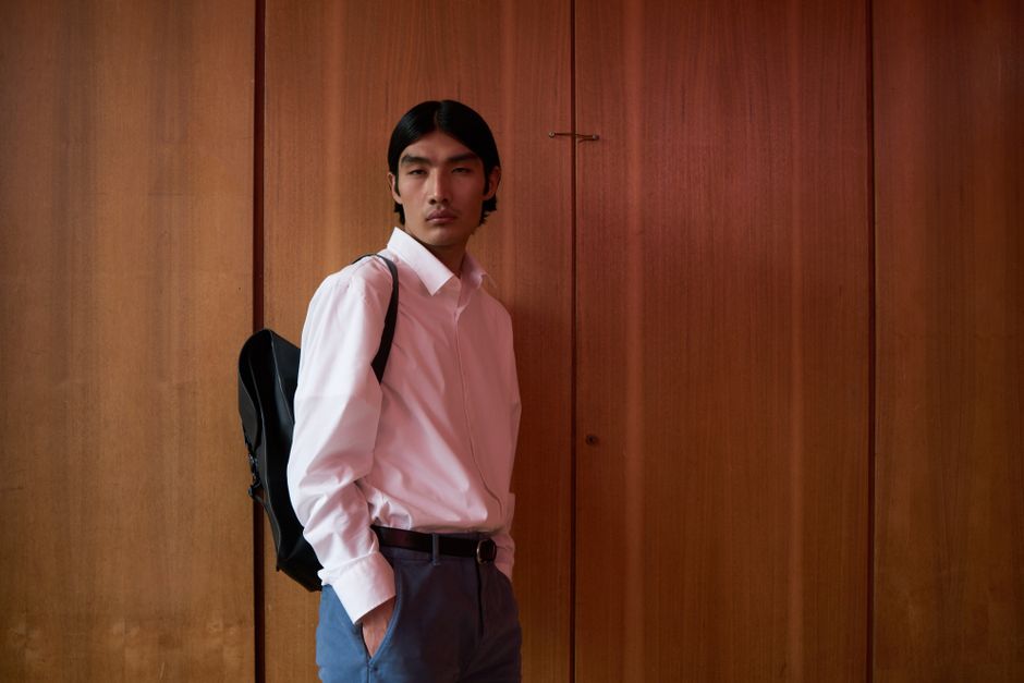 Portrait of a young man in white shirt and backpack standing against a wooden wall.