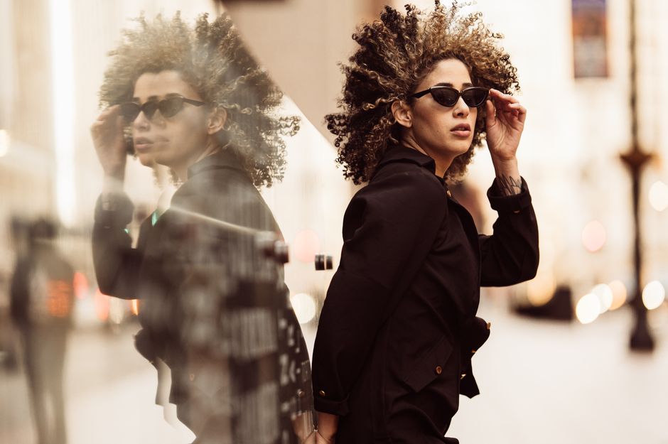 Stylish woman in a black jacket poses with sunglasses in Atlanta's urban environment with reflective glass.