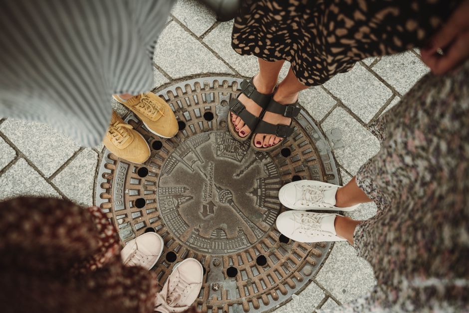 Top view of friends' shoes and sandals around a Berlin manhole cover, symbolizing unity.