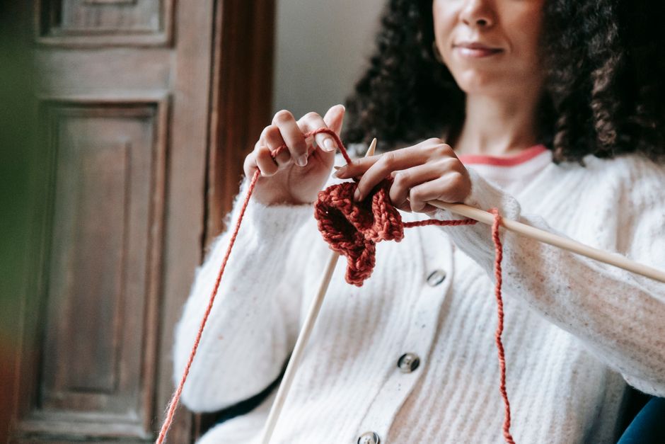 Crop unrecognizable ethnic female with curly dark hair in warm cardigan making stitches while knitting with wool yarn and wooden needles during weekend at home
