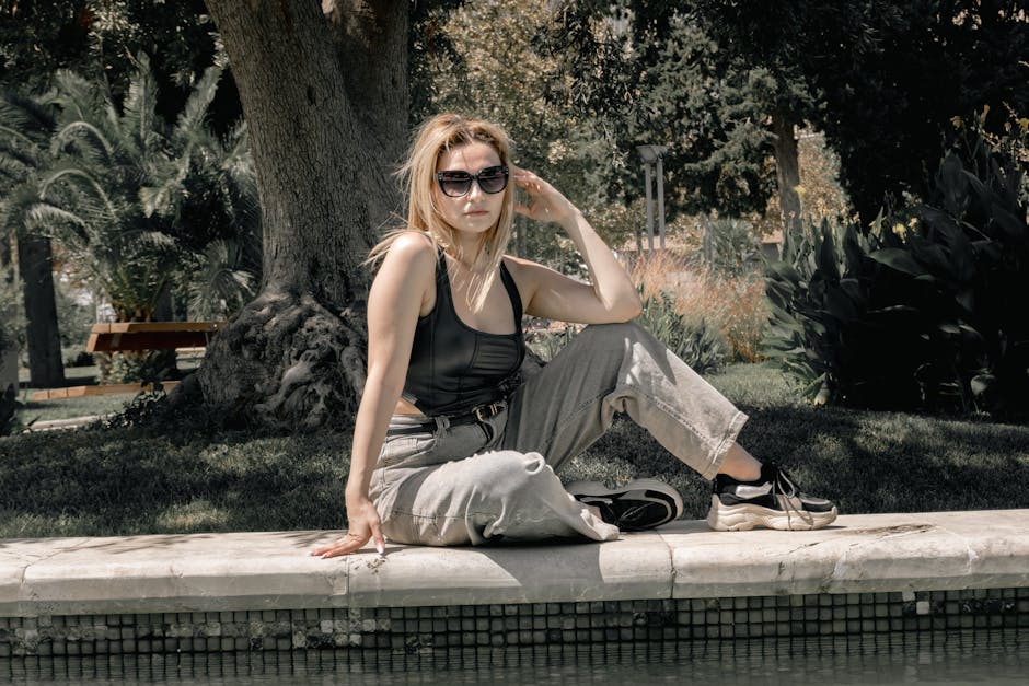 Fashionable woman sitting by a poolside in a garden, wearing sunglasses and casual clothes on a sunny day.