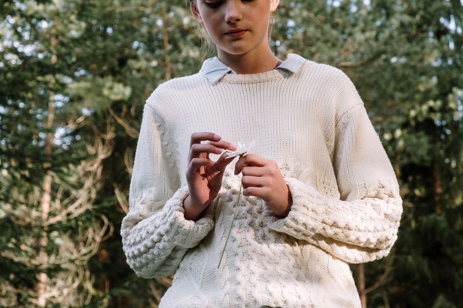 A woman in a knitted sweater holds a chamomile flower amidst a forest backdrop.
