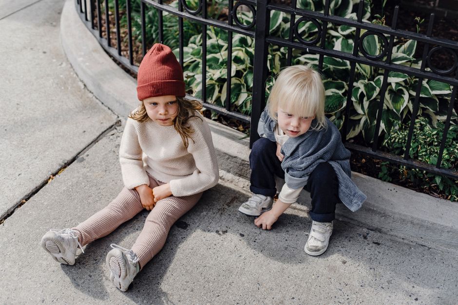 From above full body of adorable little boy and girl in hat sitting on walkway near metal fence on street