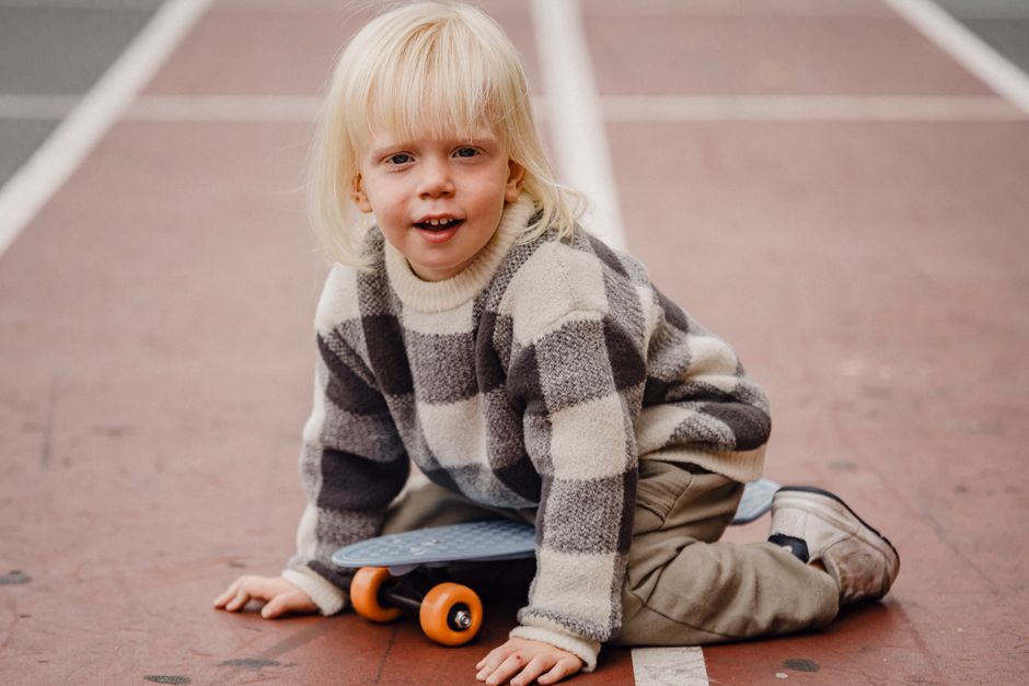 Full body of playful little kid looking at camera while sitting on sports ground with penny board against blurred background