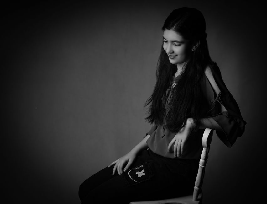 A graceful black and white portrait of a young woman sitting thoughtfully on a chair.