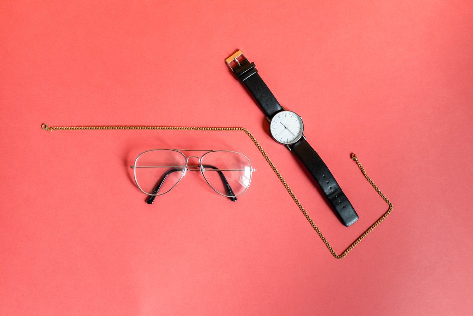 Close-up of eyeglasses, a wristwatch, and a gold necklace on a pink background.