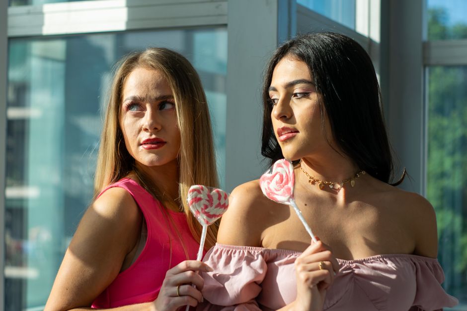 Two women enjoying pink heart-shaped lollipops in sunlight by a window.