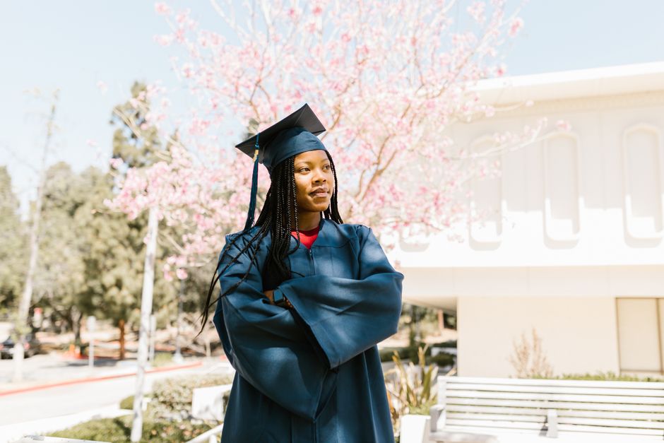 Young graduate with cap and gown stands outdoors among blooming trees, symbolizing achievement.
