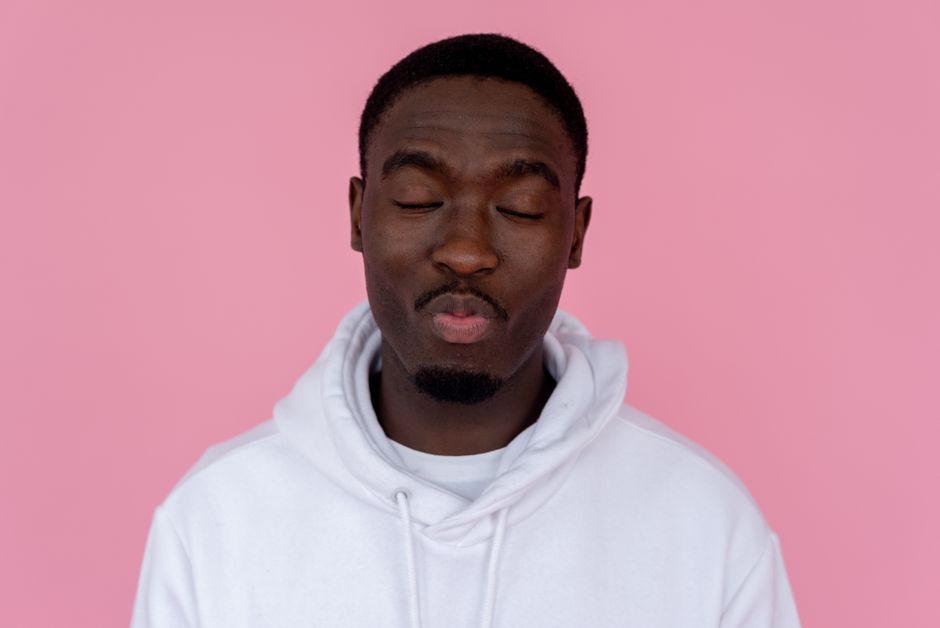 Positive African American male wearing casual sweatshirt pouting lips with closed eyes against pink background