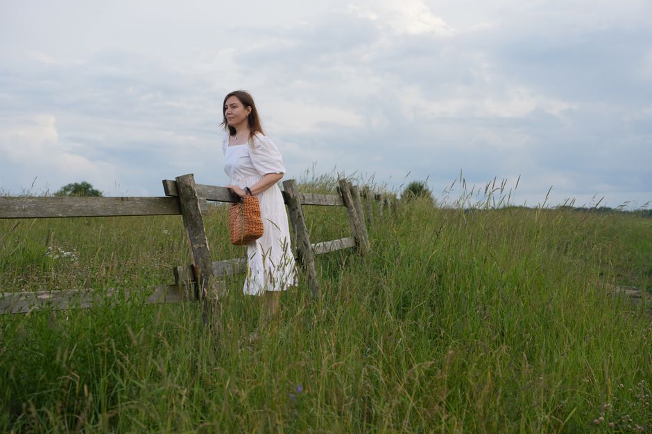 A woman in a white dress strolls along a rustic fence in a grassy field under a cloudy sky.