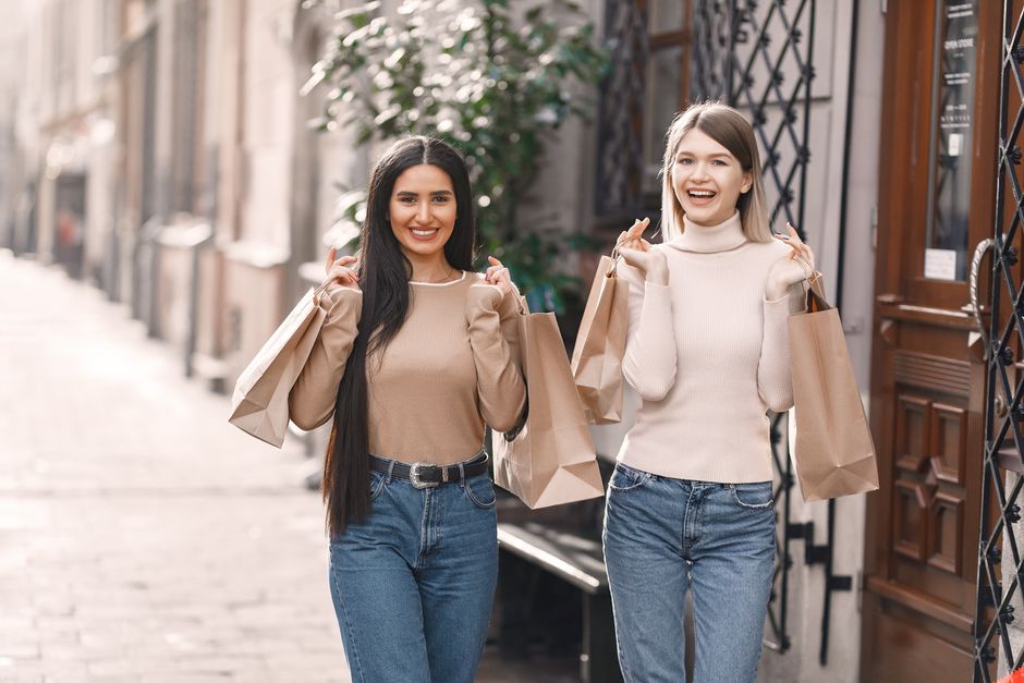 Cheerful female friends in casual outfit carrying paper shopping bags and toothy smiling at camera while walking on street