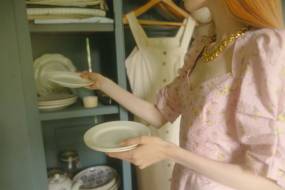 Close-up of a woman stacking plates in a kitchen cabinet, dressed in a floral pink dress.