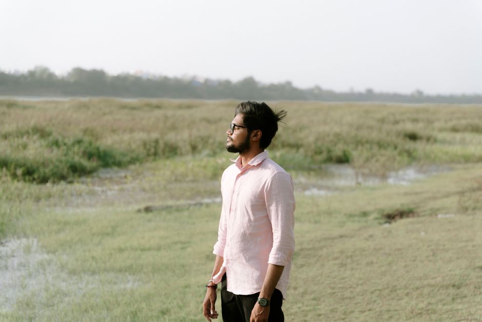 A young man in a pink shirt stands thoughtfully in a vast grassy field, looking into the distance.