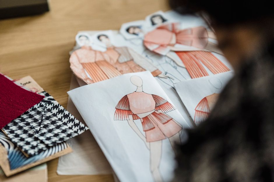 From above of crop unrecognizable female illustrator at desk with apparel sketches and ornamental fabric samples