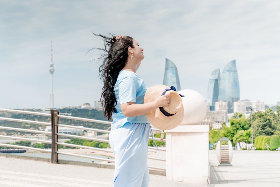 Adult woman holding a straw hat with Baku's iconic skyline in the background on a sunny day.