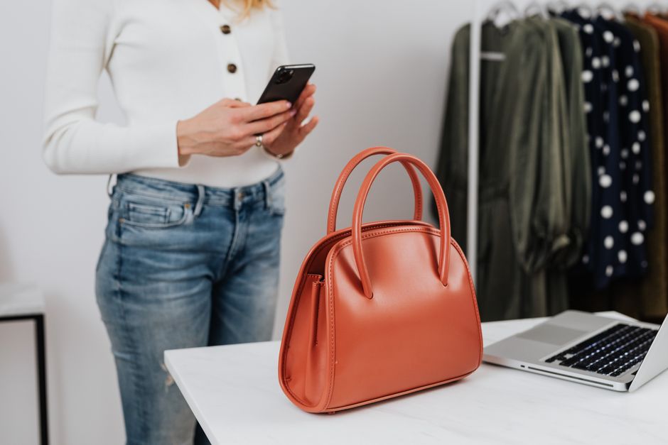 Woman in white long sleeves using smartphone in fashion studio with handbag on table.