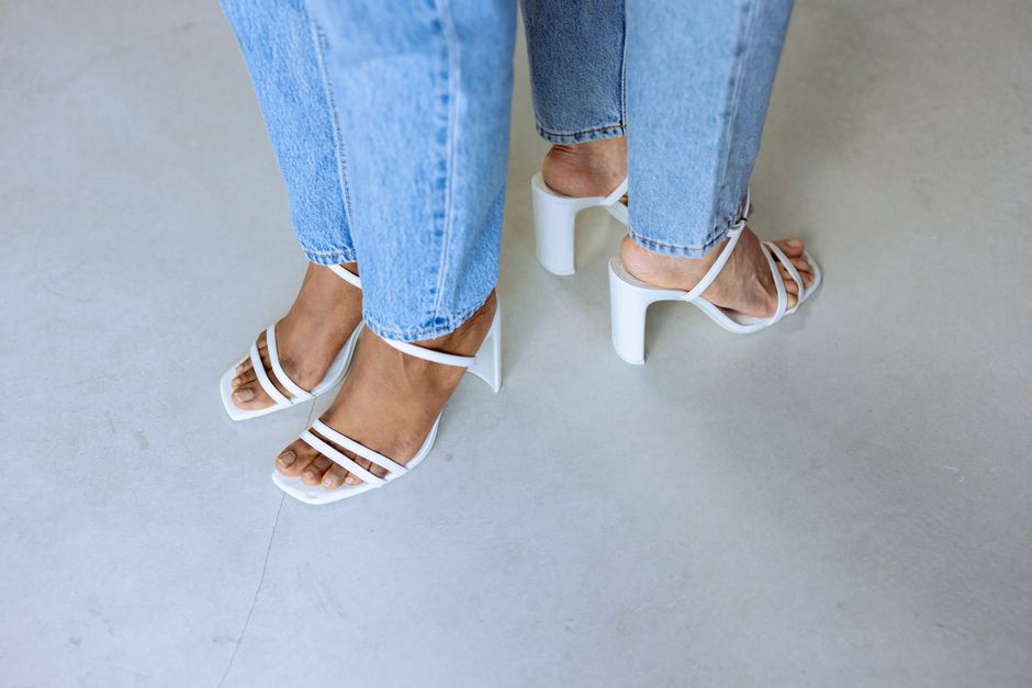 Close-up of denim-clad legs in chic white high heels on a neutral floor.