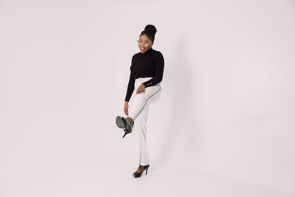 Fashionable woman in black top and white pants posing with a smile, leg raised, on a white backdrop.