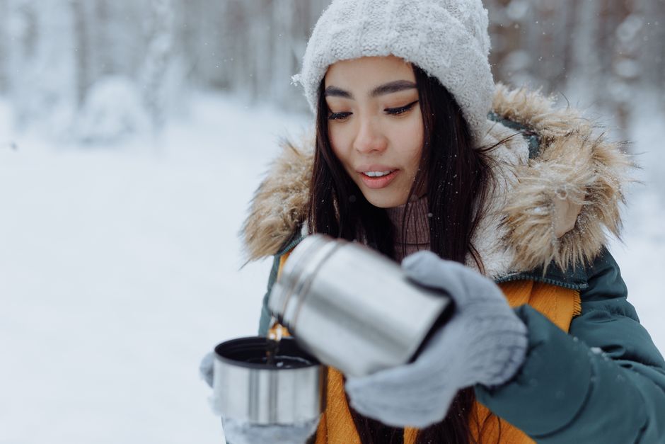 Asian woman in winter jacket pouring hot drink outside in snowy forest.