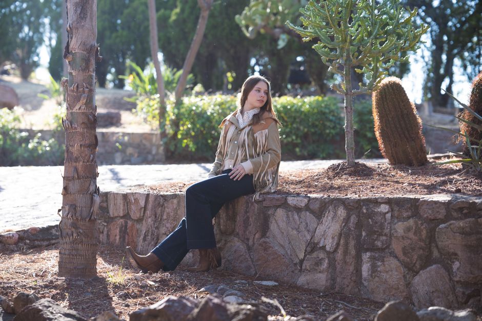 A woman in casual attire sits on a stone wall outdoors, surrounded by greenery and cacti.