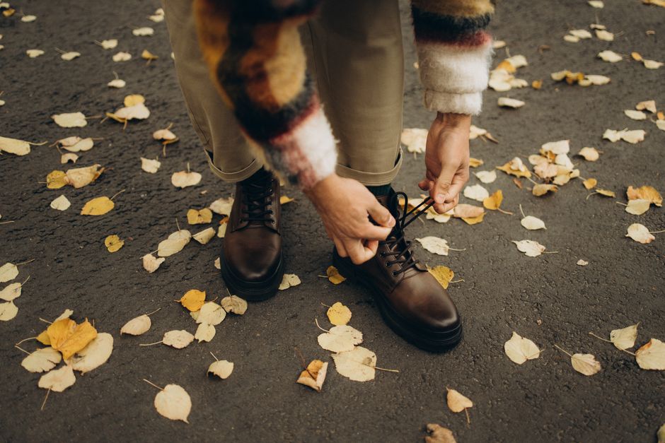 Close-up of a person tying leather boots surrounded by autumn leaves on the ground.